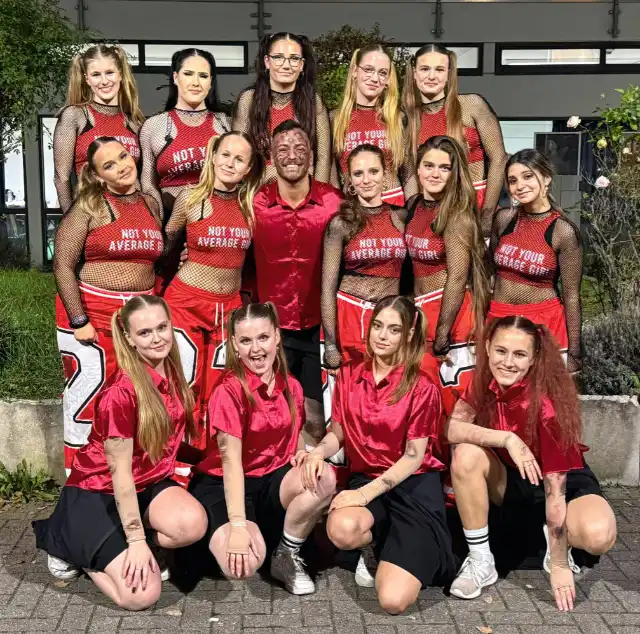 The Move, eine Gruppe von jungen Tänzerinnen posieren mit ihrem Trainer, in drei Reihen hintereinander, mit ihren roten und rot-schwarzen Tanzkostümen.